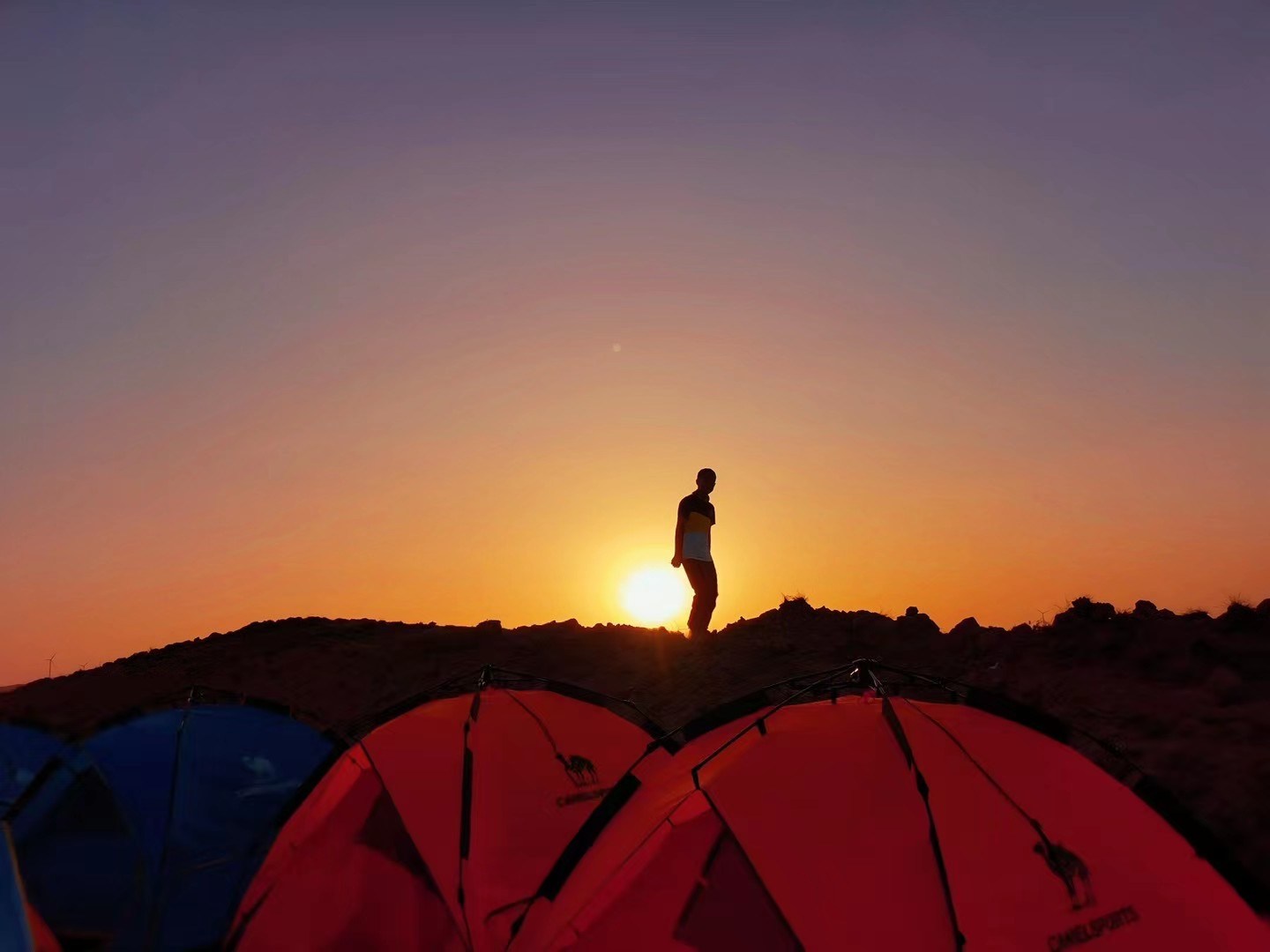 A child stands on a desert ridge as the sun rises behind a row of camping tents.