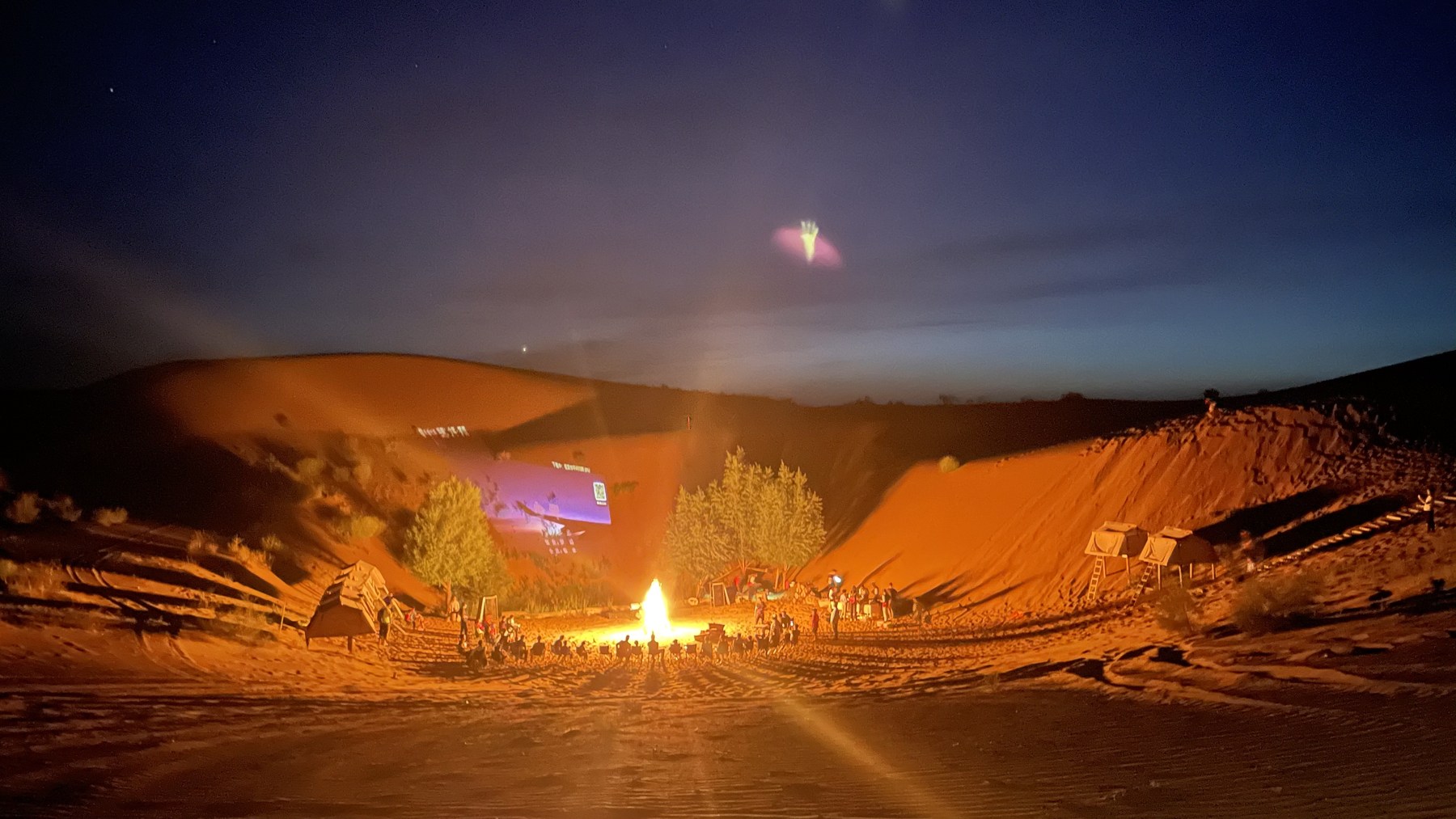 Camp participants gather around a bright bonfire between desert slopes at night.