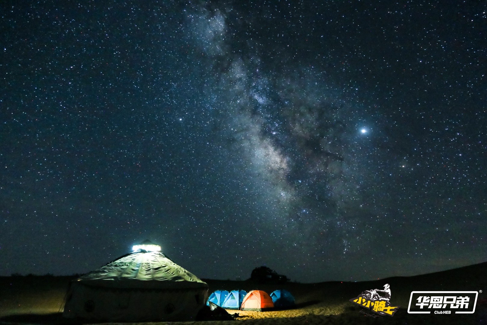 The Milky Way appears above illuminated tents in a dark desert camp.