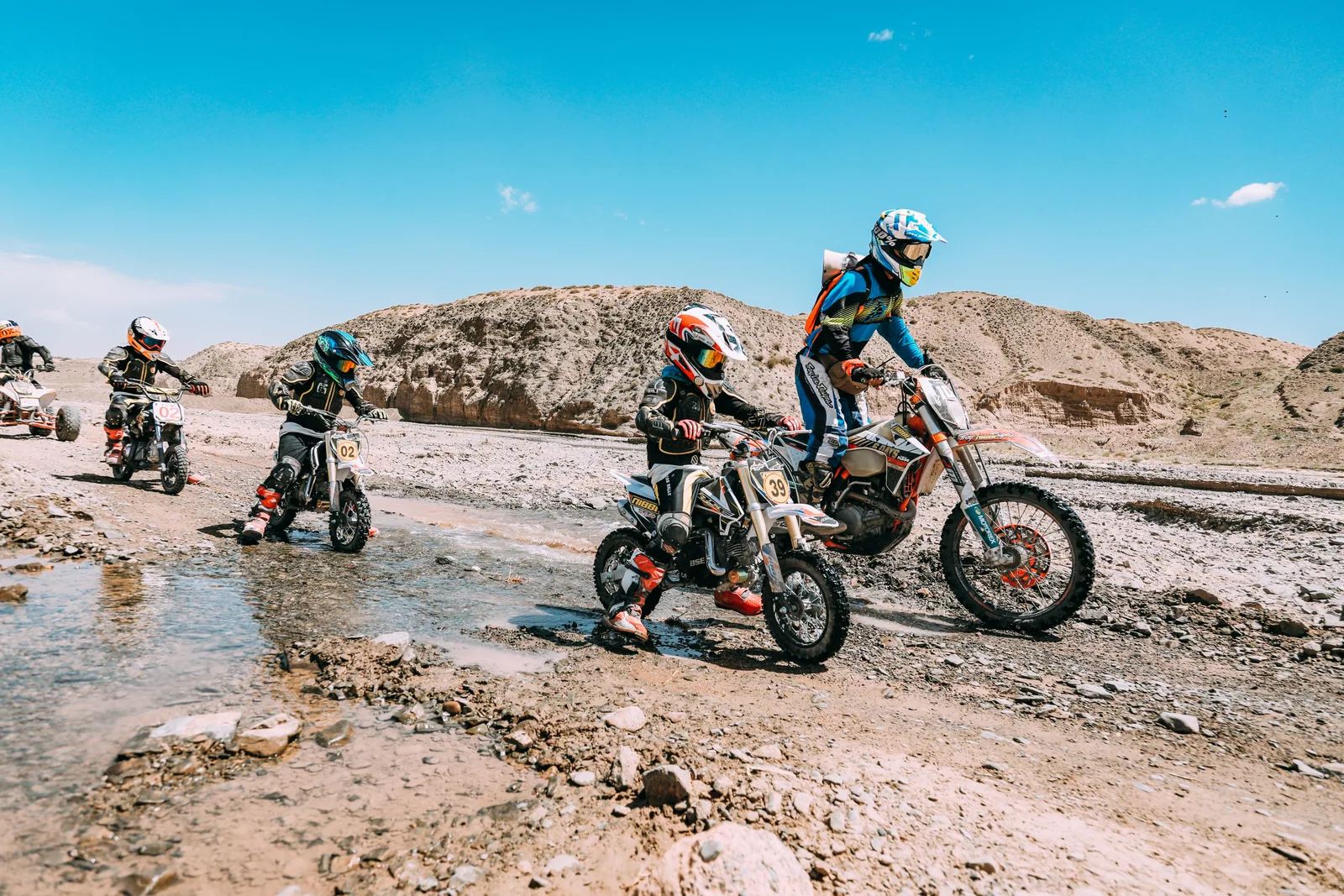 A coach accompanies young riders as they cross a shallow stream and rocky terrain