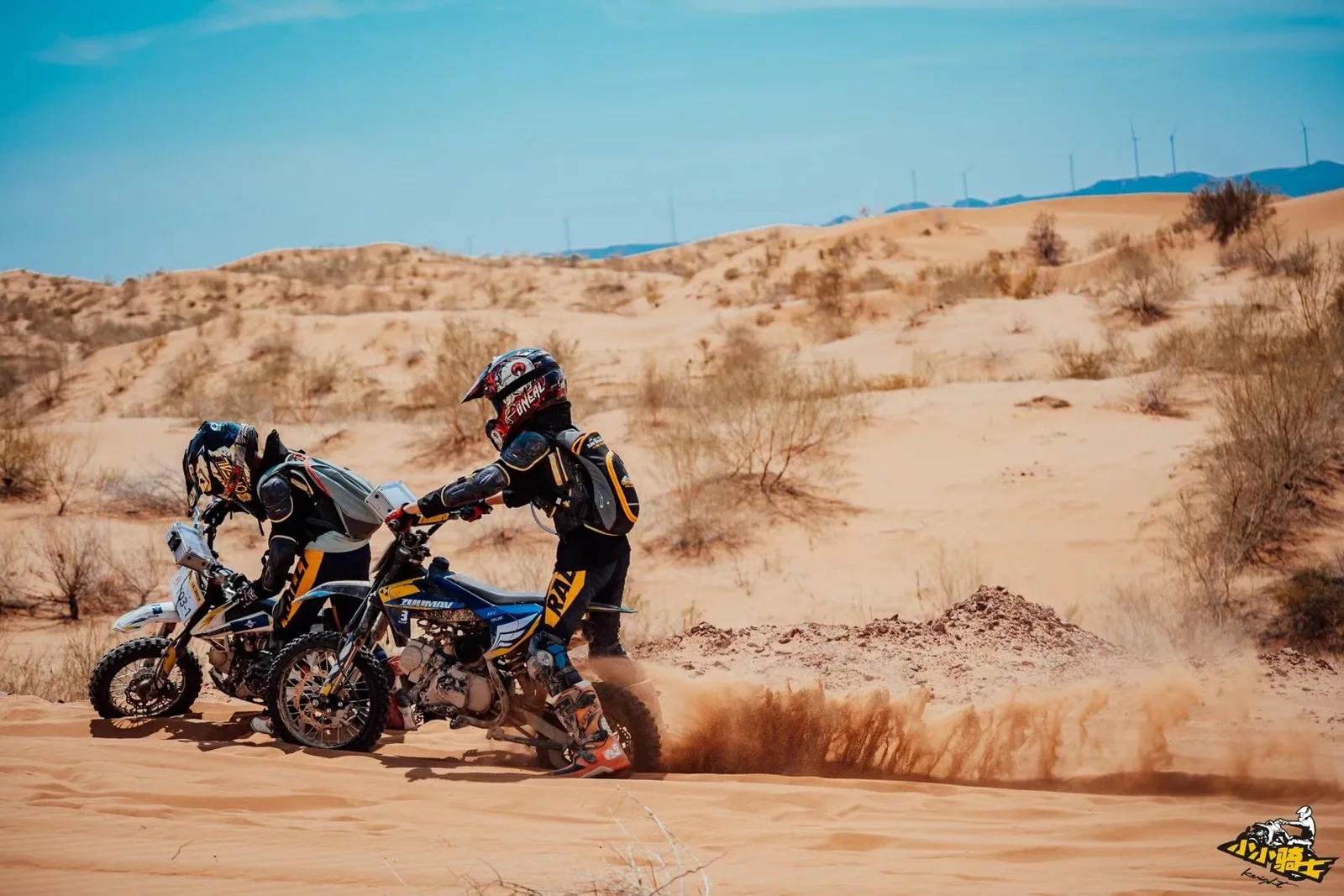 A young rider helps a teammate move a motorcycle through soft sand